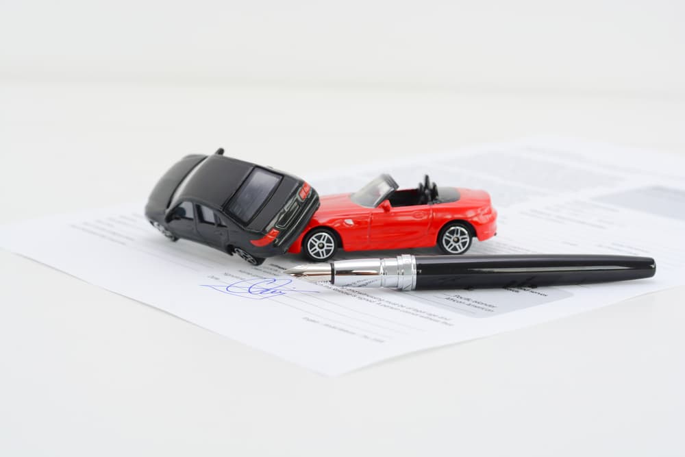 Two toy cars positioned to depict a collision, placed next to a visible insurance policy contract, symbolizing car accident damage and insurance coverage.